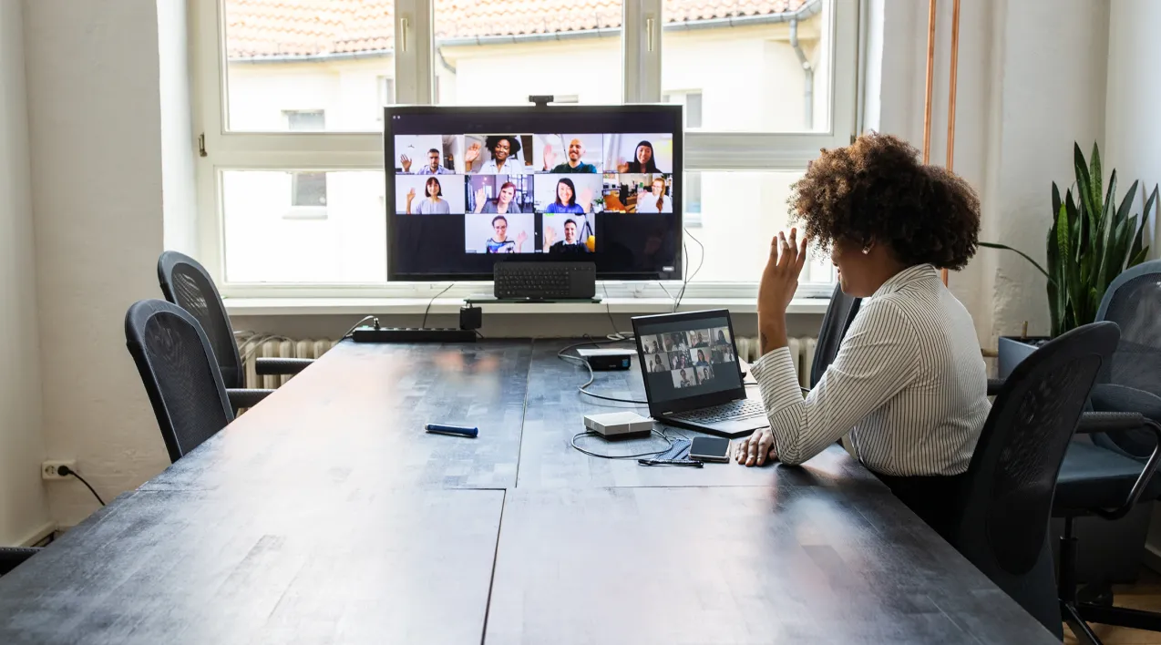 A woman sits at a conference table and waves to a large group on-screen during a digital meeting.