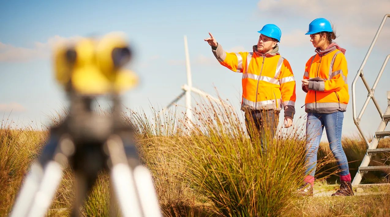 A man and a woman in high-visibility gear work on a site with a wind mill in the background.