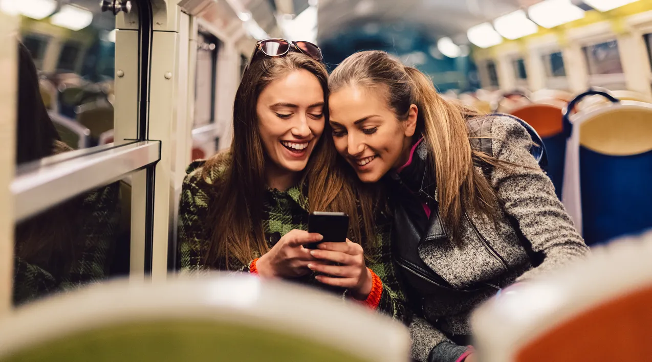 Two women share content on a mobile phone while riding a train.