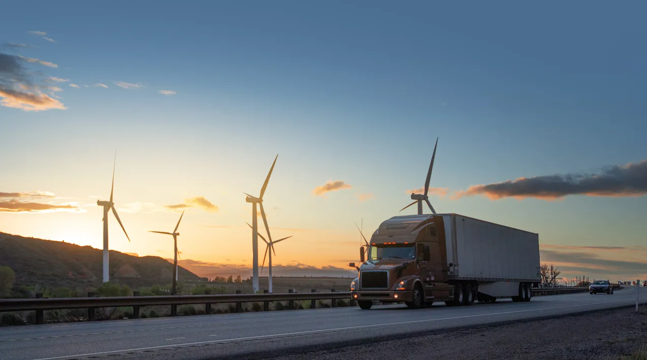 A freight truck drives on an open freeway passing by wind turbines.